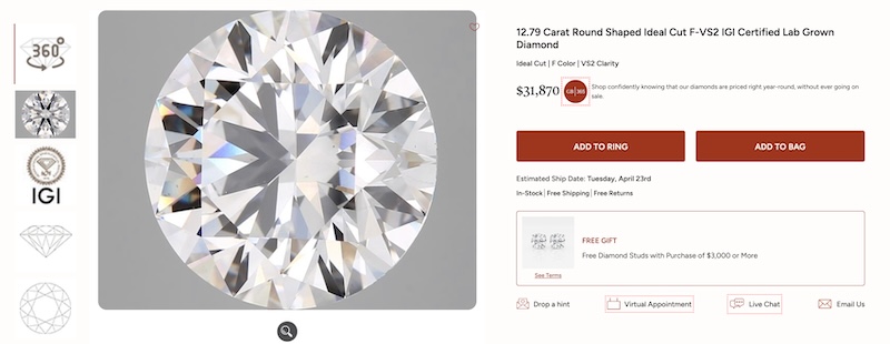 A close-up image of a 12.79 Carat Round Shaped Ideal Cut F-VS2 IGI Certified Lab Grown Diamond from Grown Brilliance. The large, sparkling diamond is displayed prominently against a gray background, with a magnifying glass icon indicating the ability to view the gem in greater detail. Beside the image, details include the price of $31,870 and buttons for 'ADD TO RING' and 'ADD TO BAG'. Additional offers such as free shipping, free returns, and a free gift with purchase are also highlighted.