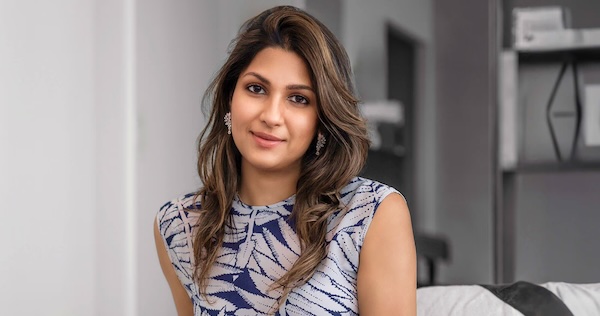 Portrait of Akshie Jhaveri, the founder of Grown Brilliance, sitting confidently in a modern room. She has shoulder-length, styled hair, and is wearing a sleeveless, patterned blue and white dress. Her expression is welcoming and her posture indicates professionalism.