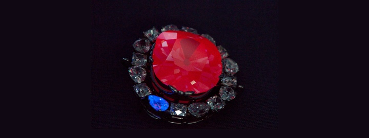 A large, vivid red gemstone, surrounded by a halo of smaller clear and blue stones, displayed against a dark background.